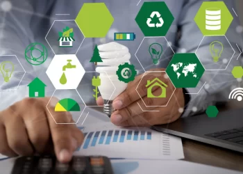 A person uses a calculator and laptop on a desk with documents. Around them float hexagonal icons symbolizing renewable energy, recycling, and sustainability, including a light bulb, wind turbines, a globe, and a solar panel—highlighting efforts to reduce their carbon footprint. | FintechZoom