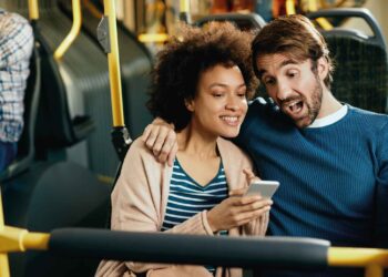 A smiling couple sits on a bus, with one person's arm around the other. They are looking at a smartphone and seem excited and happy, perhaps checking out mobile gambling in Brazil. The bus has yellow poles and a few other passengers are visible in the background. | FintechZoom