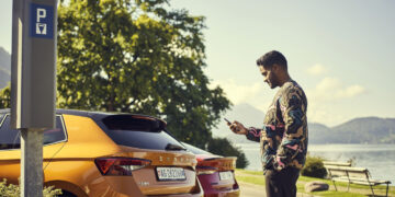 A man dressed in a colorful sweater stands next to an orange Skoda car, looking at his phone. He's in a parking area by a scenic lake with trees and mountains in the background. A parking sign and another parked car are visible nearby. | FintechZoom