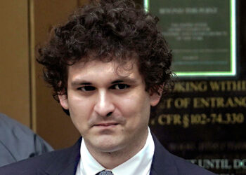 A man with curly hair, dressed in a suit and tie, stands in front of a blurred background featuring a building entrance and a sign. He appears to be outdoors, resembling Sam Bankman-Fried, with another person partially visible behind him to the left. | FintechZoom