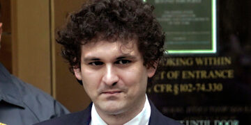 A man with curly hair, dressed in a suit and tie, stands in front of a blurred background featuring a building entrance and a sign. He appears to be outdoors, resembling Sam Bankman-Fried, with another person partially visible behind him to the left. | FintechZoom