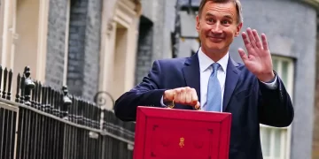A man in a suit waves with his right hand while holding a red briefcase adorned with a gold emblem, possibly containing British ISA documents, in his left. He stands outside a building with black railings and stone walls. | FintechZoom