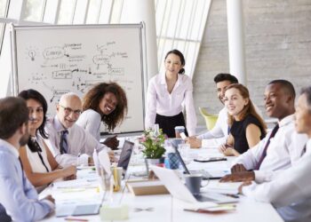 A diverse group of professionals are in a meeting room, sitting around a large table with laptops and documents. A woman in a white shirt stands and smiles at the group, discussing OKRs, while a whiteboard with diagrams and notes is visible in the background. | FintechZoom