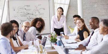 A diverse group of professionals are in a meeting room, sitting around a large table with laptops and documents. A woman in a white shirt stands and smiles at the group, discussing OKRs, while a whiteboard with diagrams and notes is visible in the background. | FintechZoom