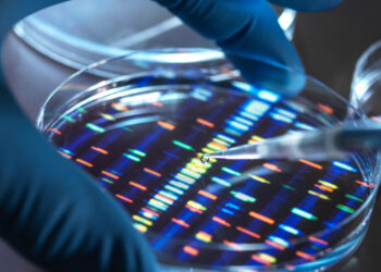 A gloved hand uses a pipette over a petri dish displaying a colorful pattern resembling a DNA sequence. The background contains another petri dish, both situated on a laboratory surface. The image emphasizes scientific research, genetics, and their impact on investment trends. | FintechZoom