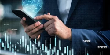 A person in a suit uses a smartphone. The background features a translucent graphic of a globe and various stock market graphs and numbers, suggesting financial data, global connections, and banking activities. The image conveys a high-tech, professional, and analytical environment. | FintechZoom