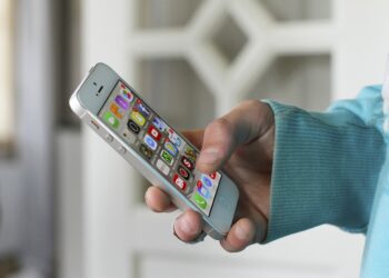 A person wearing a blue-green long-sleeve shirt holds a silver smartphone in their right hand. The smartphone display shows various colorful mobile app icons. The background is blurred, featuring a white door with a geometric design. | FintechZoom