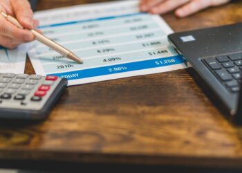 A person points at a spreadsheet with a pen, which shows various financial figures and percentages, indicating the impact of Quantitative Easing. A calculator is nearby, along with a laptop, suggesting a detailed financial analysis or budgeting task. The background is a wooden surface. | FintechZoom