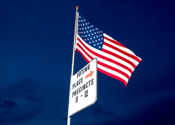 An American flag waves against a deep blue sky at dusk. Below the flag, a white sign with black text reads "VOTING PLACE PRECINCTS 11 - 12" and includes a red arrow pointing to the right, reminding citizens that their voice matters just as much as the movements in the Stock Market Today. | FintechZoom