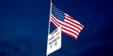 An American flag waves against a deep blue sky at dusk. Below the flag, a white sign with black text reads "VOTING PLACE PRECINCTS 11 - 12" and includes a red arrow pointing to the right, reminding citizens that their voice matters just as much as the movements in the Stock Market Today. | FintechZoom