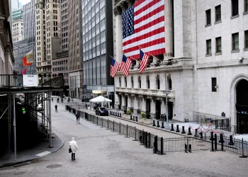 A nearly empty street in a financial district, with a large American flag hanging on a historic building. Several smaller American flags line the structure. Amidst the tall buildings and scaffolding, few pedestrians walk by, reflecting the calm before the S&P 500 trading day begins. | FintechZoom