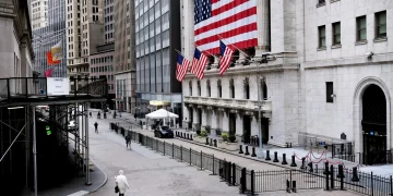 A nearly empty street in a financial district, with a large American flag hanging on a historic building. Several smaller American flags line the structure. Amidst the tall buildings and scaffolding, few pedestrians walk by, reflecting the calm before the S&P 500 trading day begins. | FintechZoom