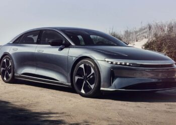A sleek, silver luxury electric sedan is parked on a sandy path near dunes and sparse vegetation. The car features a streamlined design with a distinctive continuous LED headlight strip and large black alloy wheels. The background shows a hint of a clear sky. | FintechZoom