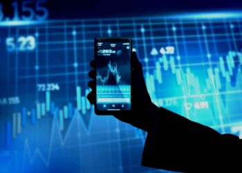 A silhouetted hand holds a smartphone displaying a stock market chart. In the background, an illuminated blue screen shows various trading graphs and numerical data, reflecting active financial information. | FintechZoom