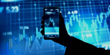 A silhouetted hand holds a smartphone displaying a stock market chart. In the background, an illuminated blue screen shows various trading graphs and numerical data, reflecting active financial information. | FintechZoom