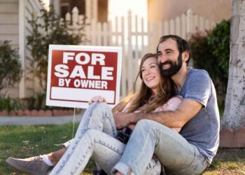 A happy couple sits on the grass, embracing and smiling in front of their new home with a "For Sale By Owner" sign. The woman is holding a sold sign, and there is greenery and a white picket fence in the background. | FintechZoom