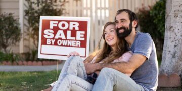 A happy couple sits on the grass, embracing and smiling in front of their new home with a "For Sale By Owner" sign. The woman is holding a sold sign, and there is greenery and a white picket fence in the background. | FintechZoom