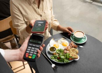 A person is making a contactless payment with a smartphone to a waiter holding a POS card reader. They are sitting at a table with a plate of food featuring fried eggs, toast, salad, and a cup of coffee. | FintechZoom