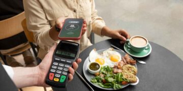 A person is making a contactless payment with a smartphone to a waiter holding a POS card reader. They are sitting at a table with a plate of food featuring fried eggs, toast, salad, and a cup of coffee. | FintechZoom