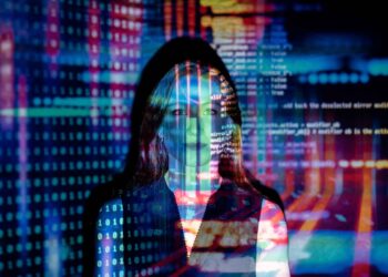 A person stands in front of a computer screen displaying colorful lines of software code and digital data. The projection of the code covers their face and body, creating a visually striking contrast between the human figure and the technological background. | FintechZoom