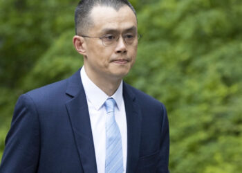 A man with short dark hair and glasses, dressed in a dark blue suit, white shirt, and light blue striped tie, is walking outside with green foliage in the background. Among his accessories is a discreet Binance pin on his lapel. | FintechZoom