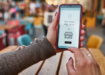 A person holds a smartphone displaying a "Buy Now Pay Later (BNPL)" option on the screen. The user is about to tap the "Buy Now" button. The scene is set in a casual outdoor setting with blurred colorful chairs and tables in the background. | FintechZoom