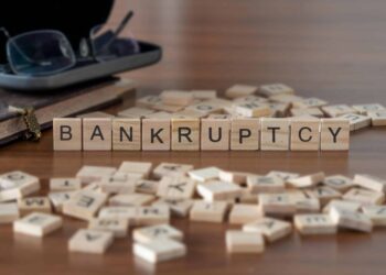 Wooden letter tiles spell out "BANKRUPTCY" on a table surrounded by scattered tiles. Nearby, a pair of glasses rests beside an old book with a ribbon bookmark, hinting at the unresolved debt in Florida that's yet to be addressed. | FintechZoom