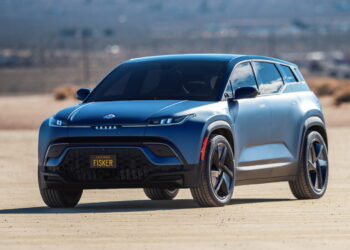 A sleek blue Fisker Ocean electric SUV is parked on a sandy terrain with a blurred, vast desert landscape in the background. The vehicle, perfect for those considering buying EV, features a modern design with a black grille, large wheels, and a license plate that reads "FISKER. | FintechZoom