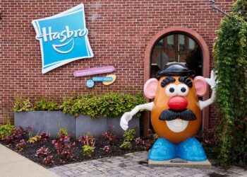 A large statue of Mr. Potato Head stands beside a building with a Hasbro sign on it. The red brick facade is adorned with green foliage and flowers at the base. The sign reads “Hasbro Worldwide Headquarters,” 101 Newport Avenue, reflecting the strength behind Hasbro stock in its vibrant presentation. | FintechZoom