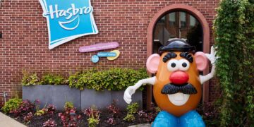 A large statue of Mr. Potato Head stands beside a building with a Hasbro sign on it. The red brick facade is adorned with green foliage and flowers at the base. The sign reads “Hasbro Worldwide Headquarters,” 101 Newport Avenue, reflecting the strength behind Hasbro stock in its vibrant presentation. | FintechZoom