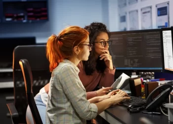 Two women sitting at a desk in an office, working on computer screens displaying code. One woman with red hair types on the keyboard, while the other with curly hair and glasses holds a tablet, thoughtfully reading a Stellar Repair for QuickBooks review. The background includes shelves and whiteboards. | FintechZoom