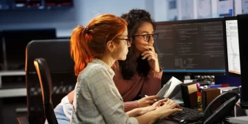 Two women sitting at a desk in an office, working on computer screens displaying code. One woman with red hair types on the keyboard, while the other with curly hair and glasses holds a tablet, thoughtfully reading a Stellar Repair for QuickBooks review. The background includes shelves and whiteboards. | FintechZoom