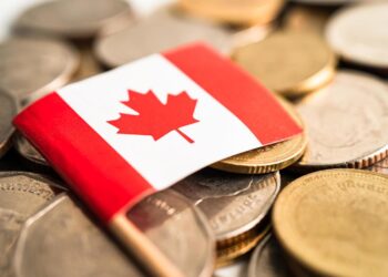 Close-up of a small Canadian flag placed on a pile of various coins, symbolizing financial diversity. The coins, reminiscent of personal loans in their variety, are different sizes and colors, suggesting a mix of currencies. The flag features the red maple leaf emblem on a white background with red bars on both sides. | FintechZoom