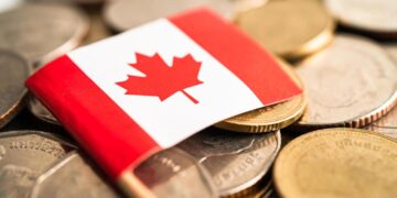 Close-up of a small Canadian flag placed on a pile of various coins, symbolizing financial diversity. The coins, reminiscent of personal loans in their variety, are different sizes and colors, suggesting a mix of currencies. The flag features the red maple leaf emblem on a white background with red bars on both sides. | FintechZoom