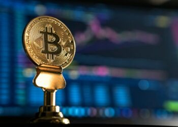 A physical Bitcoin coin displayed on a stand, with a blurred background showing colorful financial charts and graphs on a computer screen. The gold coin, often used in crypto gaming, features the Bitcoin symbol in its center. | FintechZoom