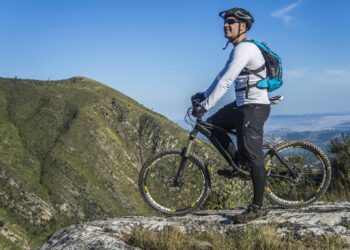 A cyclist, equipped with adaptive eyewear solutions, wearing a helmet and sunglasses, dressed in a long-sleeve shirt, shorts, and a backpack, stands with his mountain bike on a rocky ledge overlooking a mountainous landscape under a clear blue sky. | FintechZoom