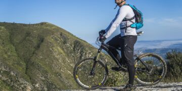 A cyclist, equipped with adaptive eyewear solutions, wearing a helmet and sunglasses, dressed in a long-sleeve shirt, shorts, and a backpack, stands with his mountain bike on a rocky ledge overlooking a mountainous landscape under a clear blue sky. | FintechZoom