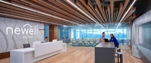 A modern office space with wooden ceiling accents and an illuminated 'Newell Brands' sign on the left wall. A person is seated at a reception desk, and two individuals are conversing at a high table on the right. Glass-walled conference rooms are visible in the background, reflecting Newell Brands' stock value in innovation. | FintechZoom
