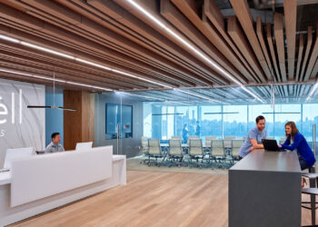 A modern office space with wooden ceiling accents and an illuminated 'Newell Brands' sign on the left wall. A person is seated at a reception desk, and two individuals are conversing at a high table on the right. Glass-walled conference rooms are visible in the background, reflecting Newell Brands' stock value in innovation. | FintechZoom