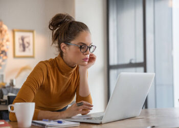 A woman with glasses and a bun is sitting at a wooden table, working on a laptop. She wears a mustard-colored turtleneck and holds a credit card, perhaps purchasing insurance. A coffee mug and stationery items are on the table. She appears to be in a modern, well-lit room. | FintechZoom