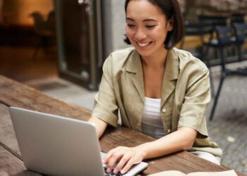 A woman with short hair, dressed in a light green shirt, is sitting at an outdoor wooden table, smiling and working on a laptop. The setting feels perfect for writing a blog post, with chairs and an open door in the background suggesting a relaxed, open-air environment. | FintechZoom