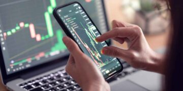 A person uses a smartphone to analyze financial charts displayed on the screen, with a laptop in the background showing a similar chart. The charts include green and red candlestick patterns, highlighting stock market data analysis to identify the best stocks to buy now. | FintechZoom