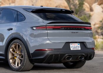 The rear view of a gray Porsche Cayenne Turbo GT, considered one of the best luxury SUVs, showcases its taillight design, quad exhaust tips, rear spoiler, and gold-colored wheels. The car is parked outdoors with rocks and greenery in the background and has a visible California license plate. | FintechZoom