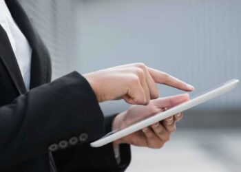 Close-up of a person using a white tablet, likely setting up digital signatures. The individual, dressed in a black suit jacket, points at the screen with one hand while holding the tablet with the other. The blurred, neutral-colored background suggests an indoor or office setting. | FintechZoom