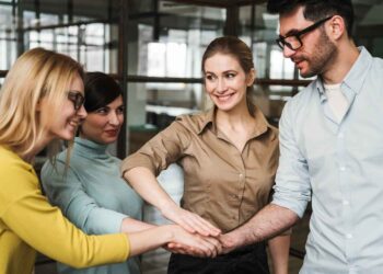 A group of four colleagues stands together in an office setting, placing their hands on top of each other in a gesture of solidarity and teamwork. They all seem to be smiling and engaged in the moment, exemplifying true collaboration. The background features glass walls and an open workspace. | FintechZoom