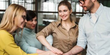 A group of four colleagues stands together in an office setting, placing their hands on top of each other in a gesture of solidarity and teamwork. They all seem to be smiling and engaged in the moment, exemplifying true collaboration. The background features glass walls and an open workspace. | FintechZoom