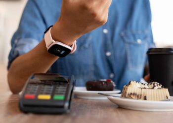 A person uses a smartwatch to make a contactless payment at a card reader terminal, seamlessly engaging in payment processing. The person is holding a black takeaway coffee cup. On the table, there are two small plates, one with a chocolate dessert and the other with a pastry. | FintechZoom