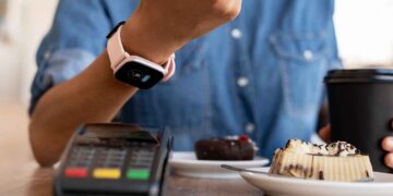 A person uses a smartwatch to make a contactless payment at a card reader terminal, seamlessly engaging in payment processing. The person is holding a black takeaway coffee cup. On the table, there are two small plates, one with a chocolate dessert and the other with a pastry. | FintechZoom