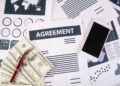 A stack of documents and charts lie on a desk, with a prominent paper titled "AGREEMENT" in bold letters. Nearby are a smartphone, a red pen, and a pile of US dollar bills, likely related to private lending. The background includes various data charts and graphs. | FintechZoom