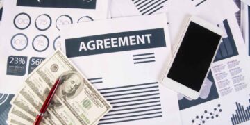 A stack of documents and charts lie on a desk, with a prominent paper titled "AGREEMENT" in bold letters. Nearby are a smartphone, a red pen, and a pile of US dollar bills, likely related to private lending. The background includes various data charts and graphs. | FintechZoom